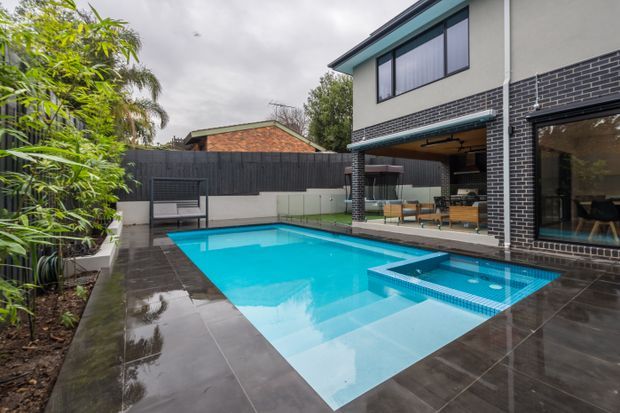 A contemporary two-storey home viewed from a landscaped backyard, centred on a bright rectangular swimming pool with an attached shallow spa or wading section. Dark large-format tiled paving surrounds the pool, with a black brick exterior wall, extensive glazing, covered outdoor seating, and a privacy fence. Tropical planting and neighbouring rooflines are visible in the background under an overcast sky.