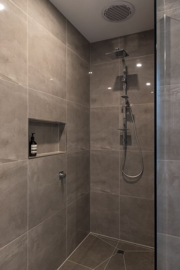 A compact contemporary walk-in shower enclosed by clear glass, finished throughout in large-format taupe-grey stone-look tiles. The shower includes a recessed wall niche with a soap dispenser, a wall-mounted rainfall and handheld shower system, a linear floor layout with central drain, recessed ceiling lights, and a circular ventilation grille. Black-framed glass edging adds contrast.