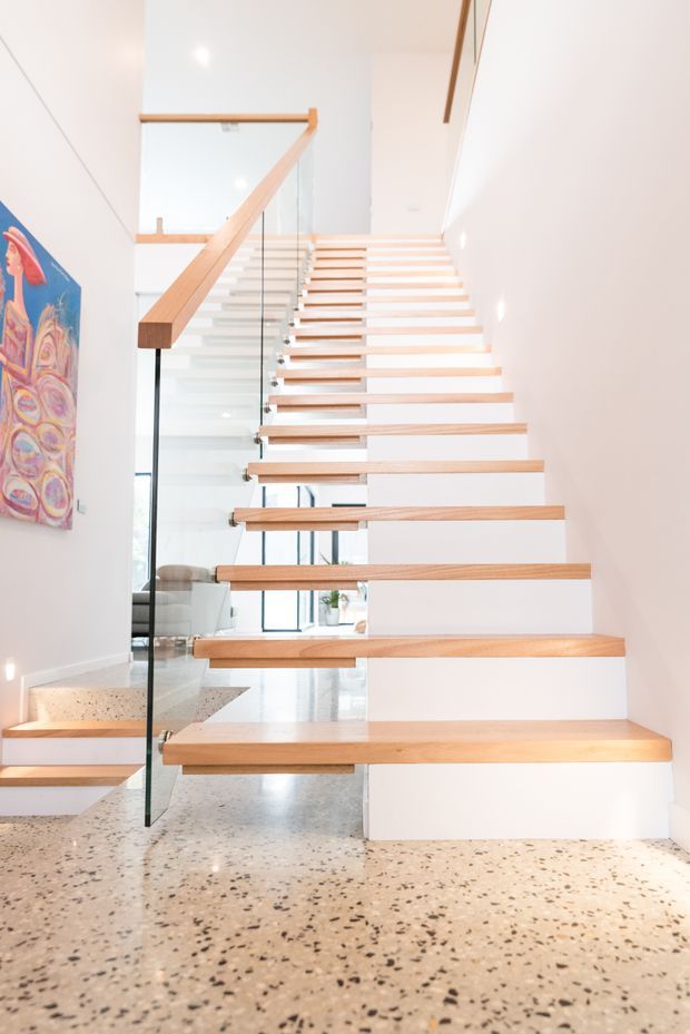Bright contemporary interior view of a floating staircase with pale timber treads, white risers and walls, and a clear glass balustrade with slim dark supports. The staircase rises beside a double-height open space, with polished terrazzo flooring and colourful wall artwork.