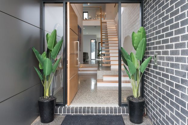 Contemporary residential entry viewed from outside, with a black brick facade and dark cladding framing a glazed timber front door. Potted tropical plants flank the entrance. Inside, a terrazzo floor leads to an open timber stair with slim horizontal treads and a visible upper-level void.