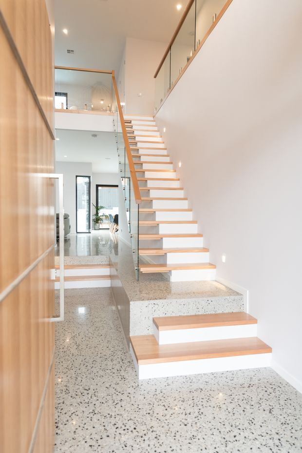 Bright contemporary multi-level interior focused on an open stairway. The staircase has pale timber treads and handrail, clear glass balustrades, and white risers, set against white walls. The floor and stair landing are finished in polished terrazzo with dark aggregate. Timber wall panelling and upper-level balustrade details are visible, with glimpses of an open living area beyond.