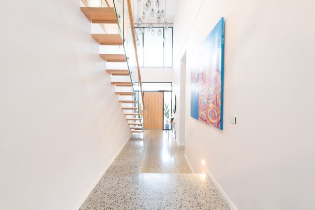 Bright minimalist hallway with polished terrazzo flooring, white walls, and a floating timber staircase with a clear glass balustrade. A tall glazed opening and timber door sit at the far end, while a colourful artwork provides a focal accent on the right wall.