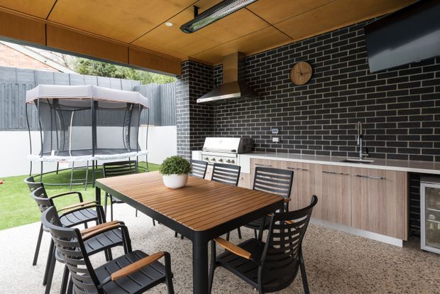 A covered outdoor entertaining area with a built-in kitchen and dining space. The room features a black brick splashback wall, timber ceiling, stainless-steel rangehood and barbecue, pale timber cabinetry, concrete-look flooring, a large timber dining table with black chairs, and a refrigerator. A trampoline and lawn are visible beyond the open side.