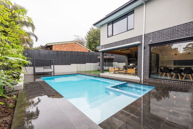 Contemporary multi-level home opening onto a landscaped courtyard with a bright rectangular swimming pool and integrated spa. The pool is surrounded by dark tiled paving, while large glazed openings reveal a dining area and an outdoor entertaining zone beneath the upper floor. Dark brickwork, pale rendered walls, timber screening, and lush planting create a private urban outdoor setting.