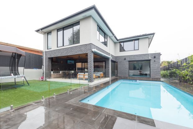 Contemporary two-storey residence photographed from the rear, featuring white rendered upper walls, dark grey brickwork, large black-framed windows, a covered outdoor entertaining area, paved pool surround and a rectangular swimming pool. A small lawn and neighbouring structures are visible around the property.