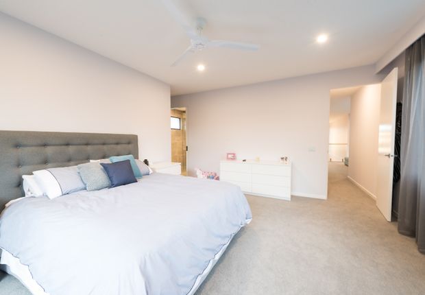 Bright, spacious contemporary bedroom with a large upholstered grey headboard, white and pale blue bedding, white dresser, recessed ceiling lights, ceiling fan, carpeted floor, and full-height curtains. A hallway or dressing area is visible through the opening at right.
