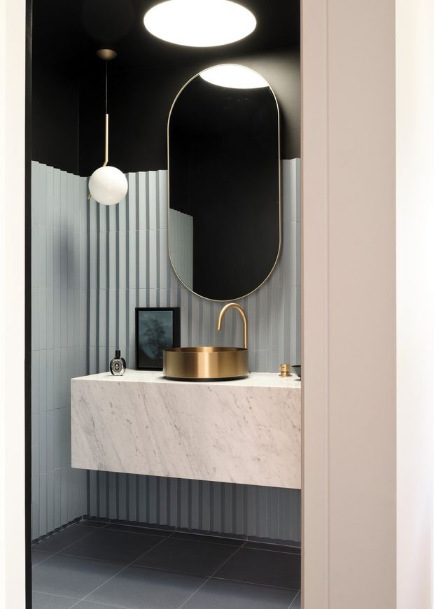 A dramatic, compact bathroom vanity viewed through a doorway. The room features a floating white marble vanity with an integrated-looking rectangular slab form, a round brushed brass vessel basin and matching curved tap, a large vertically oriented rounded-arch mirror with a slim brass frame, vertically fluted pale grey wall tiles, dark charcoal floor tiles, and a black ceiling. A spherical pendant light and recessed ceiling light provide strong illumination. The overall design is high-contrast, refined, and contemporary with brass accents.