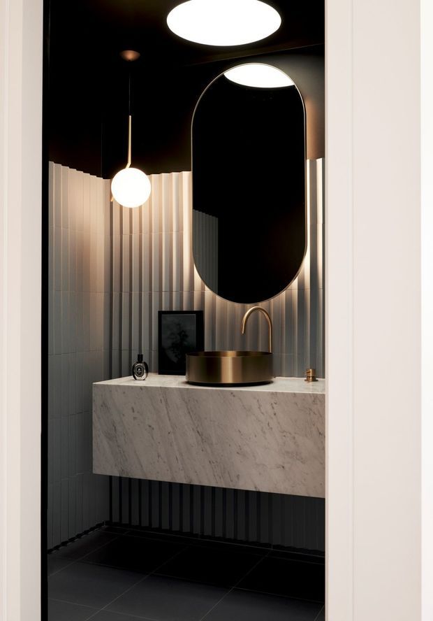 A compact, dramatically styled bathroom viewed through a doorway. The room features a floating marble vanity with a round bronze vessel basin and matching curved faucet, a large vertically oriented arched mirror, vertically ribbed wall tiles, black walls and ceiling, dark floor tiles, and sculptural globe lighting. The palette is dark, warm, and luxurious with strong geometric contrast.