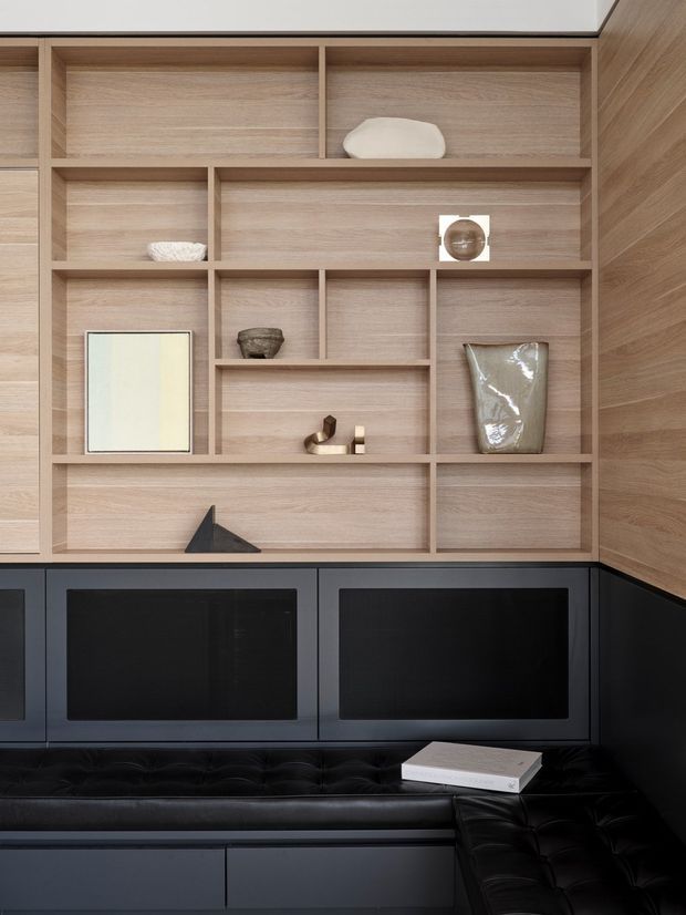 A refined built-in media and display wall with pale timber open shelving, dark lower cabinetry, integrated black screens, and a black tufted banquette wrapping the corner. Minimal decorative objects are arranged across the shelves, creating a contemporary, tailored entertainment or living-room fit-out.