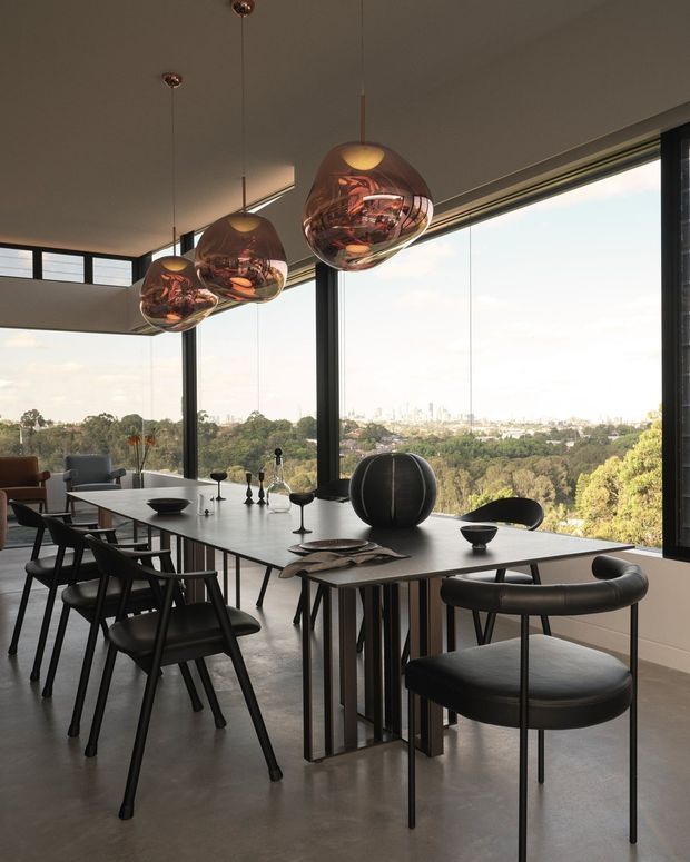 A sophisticated dining area with a long rectangular dining table positioned beside expansive floor-to-ceiling glazing. Black-framed windows overlook a leafy landscape and distant city skyline. The space features sculptural black dining chairs, a dark spherical centrepiece, and three large copper-toned pendant lights. The restrained palette of black, grey, and warm metallic accents creates a refined contemporary atmosphere.