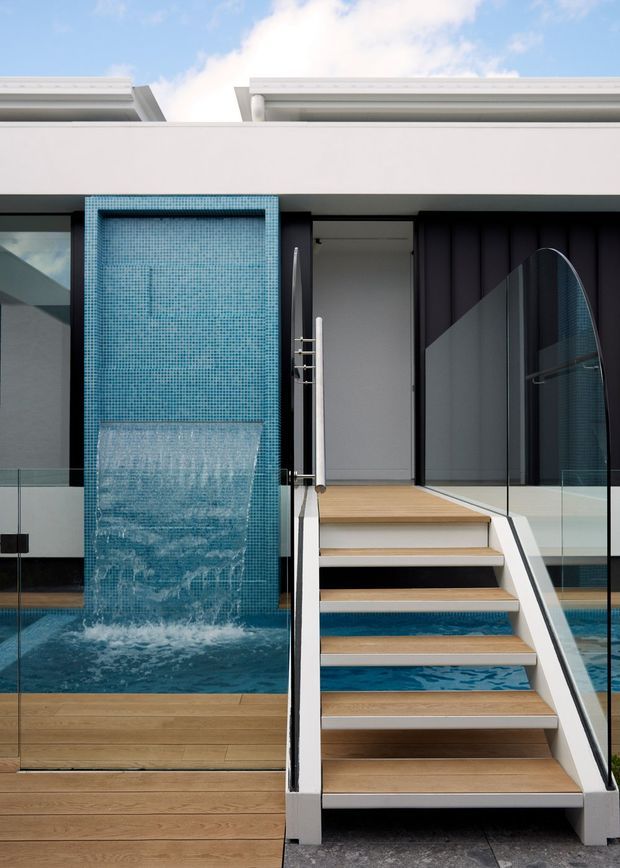 Modern residential pool courtyard featuring a turquoise mosaic-tiled water wall cascading into a swimming pool, timber decking, and a raised timber-and-white staircase with clear glass balustrades. The facade uses dark vertical cladding panels beneath a crisp white roofline, with bright daylight and clean geometric detailing.