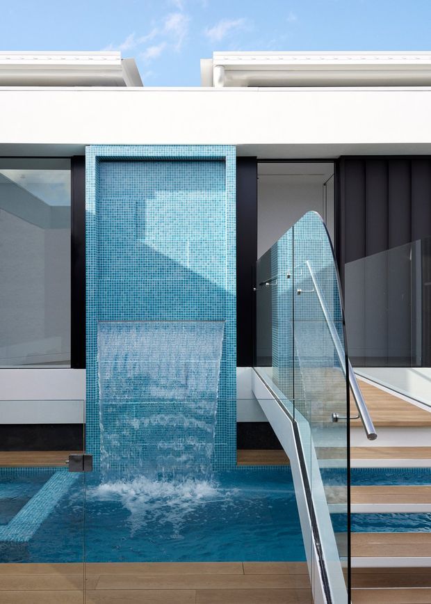A contemporary architectural pool area with a turquoise mosaic-tiled water feature cascading into a narrow swimming pool. Glass balustrades and a metal handrail border timber-toned steps, while dark vertical wall panels and white projecting roof forms create a crisp, modern composition. Bright daylight and clear blue water give the scene a clean, refreshing atmosphere.