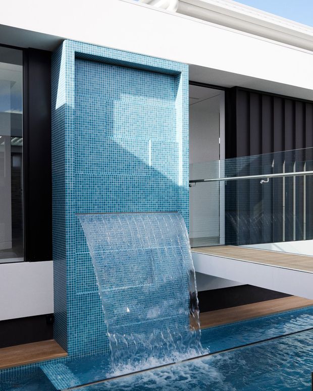 Close-up exterior view of a contemporary pool courtyard featuring a tiled turquoise mosaic water-feature wall and broad sheet waterfall flowing into a swimming pool. The surrounding building has crisp white soffits, black vertical cladding, large dark-framed glazing, and clear glass balustrades with timber decking.