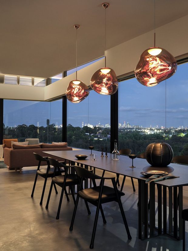 A sophisticated open-plan dining and living area at dusk, featuring a long dining table with black chairs, a lounge seating area, sculptural pendant lights, polished concrete flooring, and expansive floor-to-ceiling glazing overlooking a city skyline and treetops.