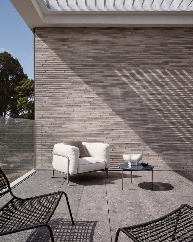 A sunlit contemporary terrace with a grey stacked-brick feature wall, pale concrete or stone paving, frameless glass balustrade, and a white pergola-like overhead structure casting strong linear shadows. Minimal outdoor furniture includes a white upholstered lounge chair, a small round black coffee table, and black woven loungers.