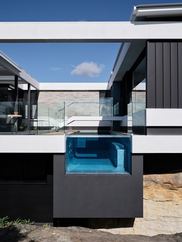 A sharp contemporary courtyard exterior centered on a compact elevated swimming pool with a transparent glass front. The home features crisp white and charcoal geometric volumes, dark vertical cladding, extensive frameless glass balustrades, a flat roofline, and a pale horizontal masonry wall. Natural rock is integrated into the lower site, while the clear blue sky and strong sunlight emphasize the minimalist architectural form.