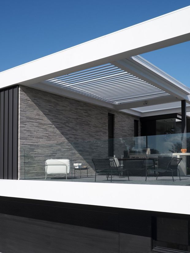 Modern residential exterior showing an elevated terrace or balcony with glass balustrades, dark outdoor furniture, a grey stacked-stone feature wall, black vertical cladding, and a white projecting roof slab. A large slatted pergola creates a covered outdoor sitting area, with the lower level partially visible beneath.