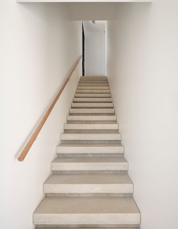 Minimalist interior stairway viewed from the bottom, with broad light grey concrete or stone treads, white walls, and a simple timber handrail along the left wall. The staircase rises toward a narrow landing or upper hallway, creating a clean, understated architectural composition.