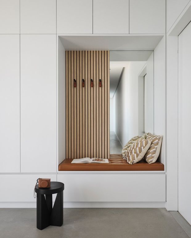 A minimalist interior hallway or entry area featuring a built-in recessed window seat with a warm timber slatted backing, leather-toned cushion, and patterned pillows. White full-height cabinetry and wall panels frame the niche, while a long corridor extends behind it. The space has a polished concrete floor and a small black side table with a leather bag in the foreground.