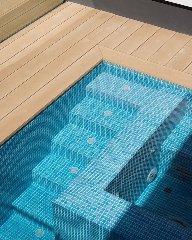 Close-up architectural photograph of a tiled swimming pool with broad submerged steps and integrated bench-like platforms. The pool is finished in small turquoise-blue mosaic tiles, with visible circular drains and pool fittings. A warm light-timber deck borders the pool, and a dark wall edge is visible in the background. The composition focuses on the pool interior and its stepped geometry.