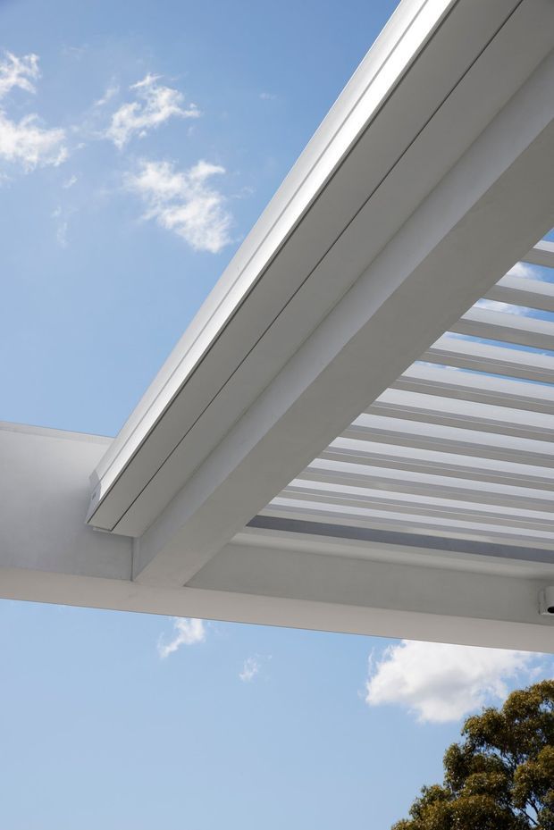 Upward-looking view of a white architectural pergola or covered outdoor structure, with broad beams and evenly spaced translucent or open slats forming a strong geometric composition against a bright blue sky. The image primarily highlights the roof structure and clean modern detailing; trees are visible only at the lower edge.