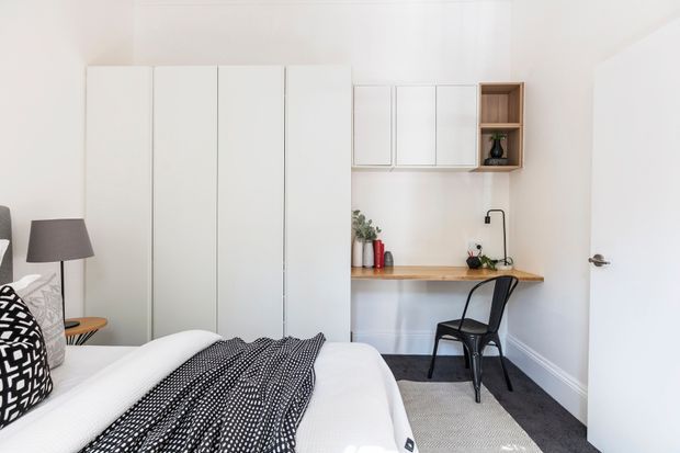 Bright, compact bedroom with a white upholstered bed in the foreground and a black-and-white patterned throw and cushion. Along the back wall is a built-in white wardrobe, while a timber-topped wall-mounted desk with a black chair sits beneath white overhead cabinetry and an open timber shelf. A small black wall-mounted tap and basin area are visible at the desk, with neutral carpeting, white walls, and simple contemporary furnishings.