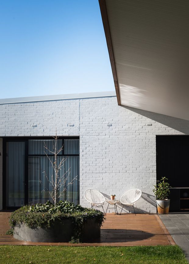 A sunlit contemporary courtyard or patio beside a white-painted brick building. The composition features a timber-lined roof overhang, dark-framed glazed doors, a black opening or doorway, timber decking, concrete pavers, raised concrete planters, outdoor lounge chairs, and established planting. The image emphasizes the relationship between the low building facade, sheltered outdoor space, and landscaped garden.