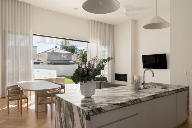 Bright contemporary open-plan kitchen and dining area with a large marble island in the foreground, pale streamlined cabinetry, a round dining table with upholstered chairs, and full-height sheer curtains framing a suburban outlook. Two sculptural pendant lights, concealed appliances, and neutral finishes create a refined, airy interior.
