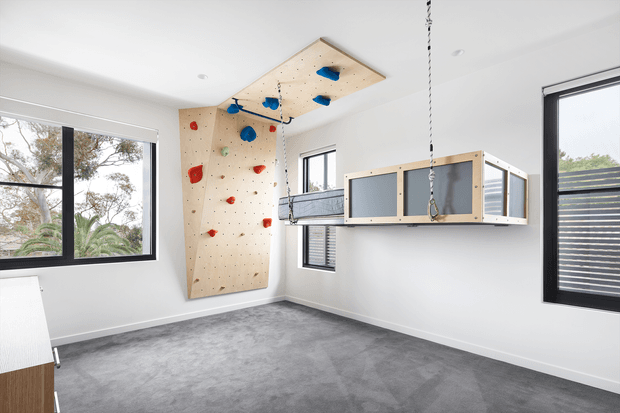Bright, spacious room with white walls, large black-framed windows and grey carpet, featuring a built-in indoor climbing wall with colourful holds, an overhead climbing panel, and a suspended wooden-framed cubby or play structure. The room appears designed as an active children's play or recreation space.