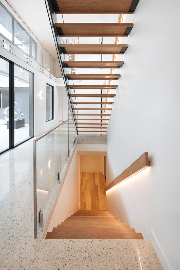 A bright contemporary interior stairwell viewed from an upper landing, with a timber staircase descending to a lower level. Floating timber ceiling slats span a tall void above, supported by dark metal brackets, while glass balustrades line the landing. The space features white walls, large black-framed glazing, polished terrazzo flooring, timber flooring below, and integrated linear lighting along the stair wall.