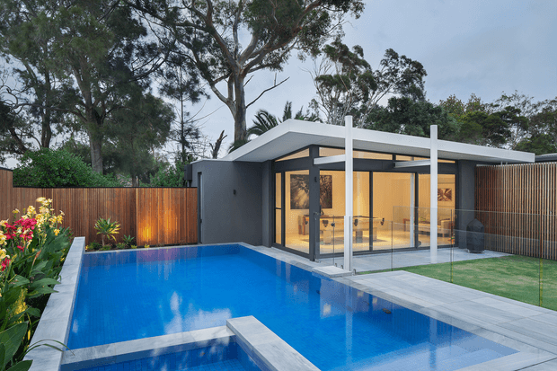 A modern single-storey residence viewed from the backyard, featuring a flat-roofed pavilion with extensive black-framed floor-to-ceiling glazing, a warm illuminated interior, and a narrow white structural frame. The home opens directly onto a vivid blue rectangular swimming pool with a raised concrete diving platform or bridge, pale paved surrounds, timber privacy fencing, landscaped planting, and lawn.