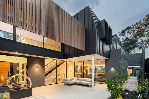 Contemporary multi-level residence viewed from a landscaped courtyard, featuring dark metal cladding, extensive floor-to-ceiling glazing, warm timber vertical screening, sharp angular roof forms, and an open-plan living area visible through the glass. The courtyard includes a pale paved terrace, outdoor seating, planting, and sculptural decor.