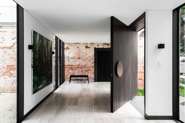 A contemporary residential entry hall with a long, light-toned tiled floor, white walls, black-framed glazing, and a dramatic oversized dark timber pivot door opening toward an exterior courtyard. Exposed original red brick walls are visible at the far end and through the glazing, contrasting with the crisp modern interior. A large dark abstract artwork, narrow bench, and minimal wall lighting complete the restrained space.