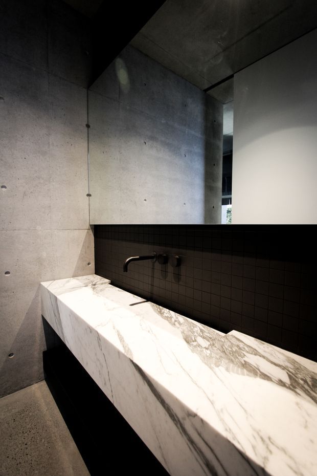 A dramatic minimalist bathroom vanity with a thick white veined stone benchtop, black under-bench cabinetry, a dark small-format tiled splashback, exposed concrete walls and ceiling, and a wall-mounted dark metal tap. A large frameless mirror spans the upper wall, reinforcing the moody, monolithic aesthetic.