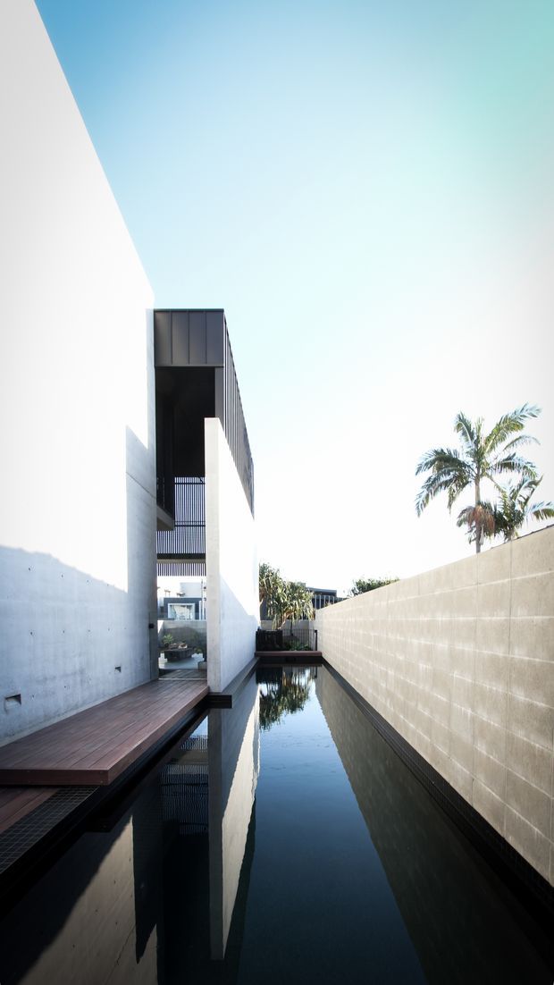 A narrow modern architectural courtyard with a long reflecting pool running between smooth white walls and a raw concrete boundary wall. Timber decking and dark metal detailing line the water, while a tall cantilevered or framed upper volume creates a dramatic vertical composition. Palm trees are visible beyond the courtyard, and the bright open sky emphasizes the minimalist design.