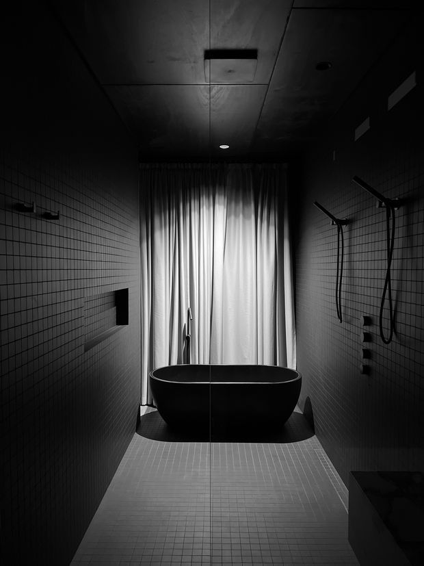 A dark, dramatic modern bathroom viewed down a narrow tiled space. A freestanding black oval bathtub sits in front of full-height pleated curtains, with wall-mounted shower fixtures and hand showers on both sides. The room has a dark ceiling with recessed lighting, small square wall and floor tiles, and strong monochrome contrast.
