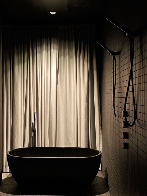 A moody, dark bathroom featuring a freestanding matte-black oval bathtub, black wall-mounted bath fixtures, a tiled wall, and full-height pleated curtains softly illuminated from behind. The space has a dramatic, minimalist spa-like atmosphere.