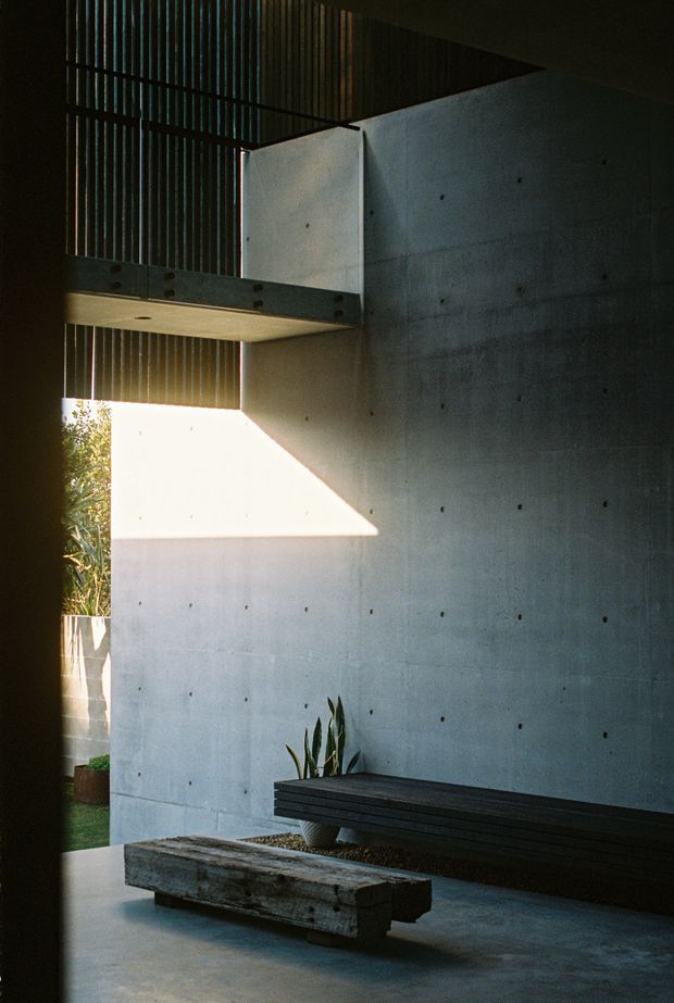 A dramatic minimalist interior courtyard or entry space with exposed board-formed concrete walls, a dark concrete floor, a heavy timber bench, and a long dark built-in bench beneath a steel or concrete mezzanine. Vertical slatted screening filters daylight from the upper level, creating strong geometric shadows and a moody, architectural atmosphere. A small potted plant and glimpses of greenery are visible at the edge.