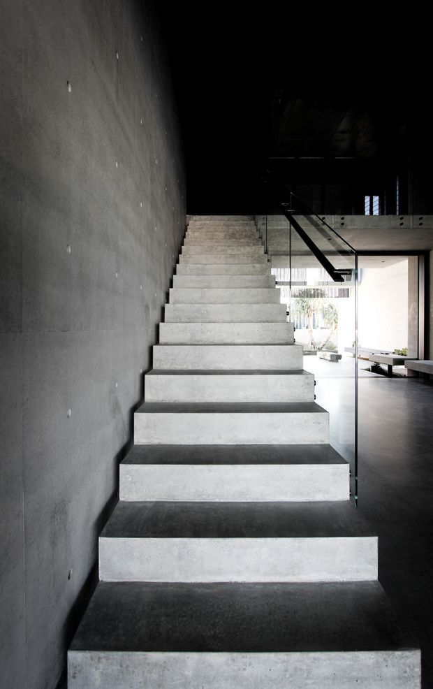 A dramatic interior stairway with broad monolithic concrete steps ascending alongside a raw concrete wall. A clear glass balustrade with dark metal handrail borders the stair, leading toward an open, double-height concrete interior with glimpses of a bright courtyard and minimal built-in seating beyond. The scene has strong contrasts between deep shadows and daylight, with an austere contemporary architectural character.