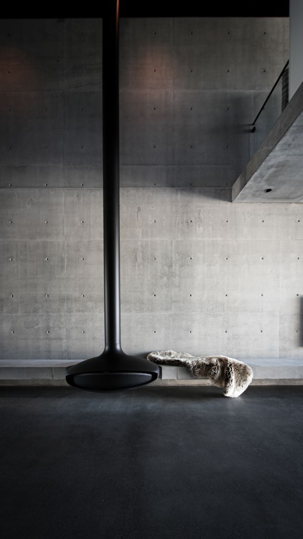 A dark, minimalist interior with exposed board-formed concrete walls and ceiling, visible construction joints, and a large suspended black cylindrical fireplace or architectural heater. A low concrete bench runs along the wall with a draped fur throw. The space has a dramatic, restrained industrial character with strong vertical and horizontal lines.