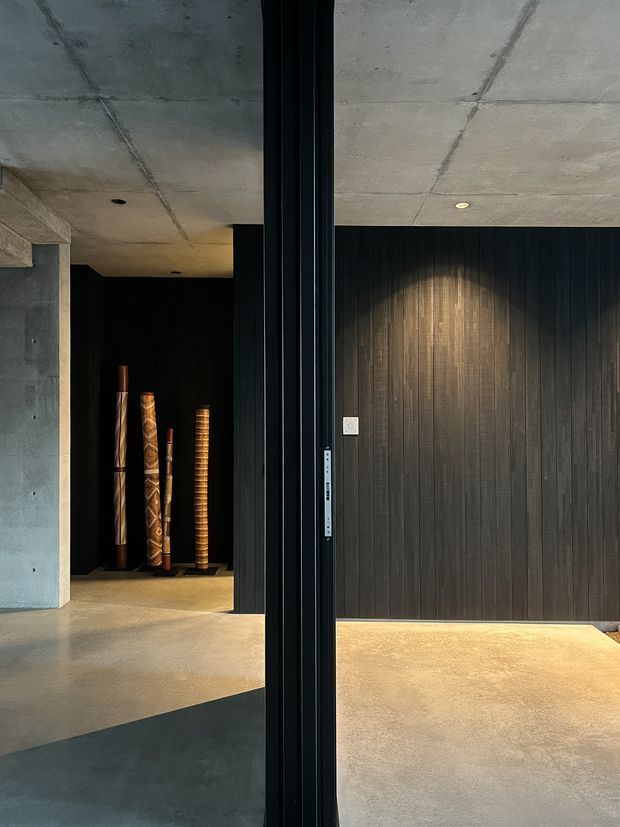 A moody contemporary interior featuring exposed concrete ceilings and walls, dark vertical timber wall panelling, polished concrete flooring, and a large black-framed glazed opening or pivot door. Decorative timber objects are displayed in a recessed niche, with concealed floor-level lighting and focused wall lighting creating a refined, dramatic atmosphere.