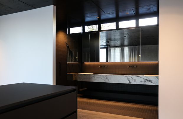 A dark, sophisticated bathroom viewed from an adjacent area, featuring a long double vanity with a dramatic white marble-look benchtop, black cabinetry, dark small-format tiled walls and floor, a wide mirror, warm integrated lighting, and high clerestory windows. A freestanding or partially visible bathtub appears at the left foreground.