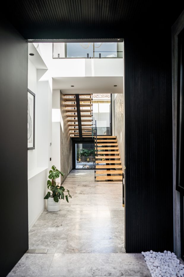 A dramatic contemporary interior entry hall with a double-height void and floating timber staircase. The space features polished light stone flooring, white plaster walls, a dark ribbed ceiling and dark vertical feature wall, extensive glazing, and a black metal-framed staircase with open timber treads. A potted plant and white decorative stones soften the otherwise minimalist architectural composition.