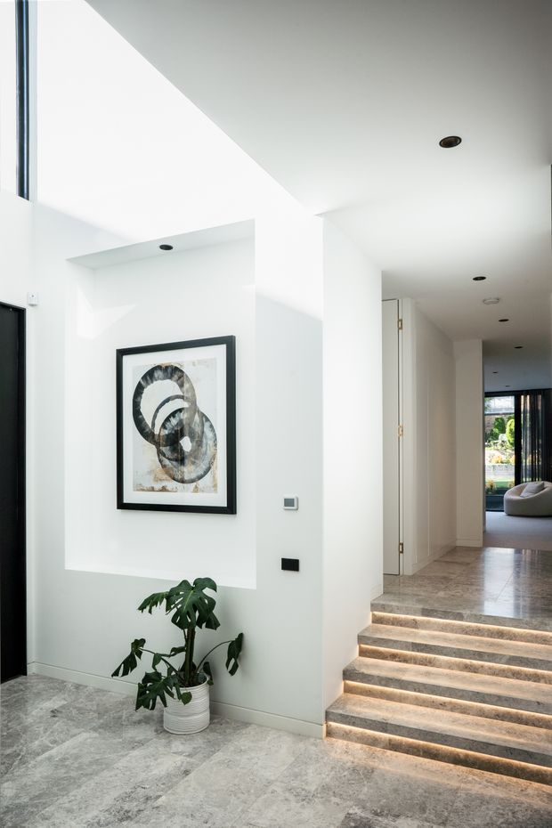 Bright contemporary interior entry hall with white walls, a recessed art display niche, framed artwork, indoor plant, and a short illuminated stone staircase leading to a corridor and living area. The space has recessed ceiling lights, pale grey stone flooring, and clean minimalist detailing.