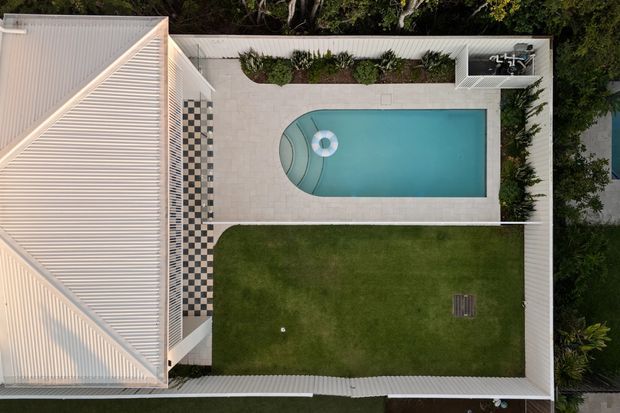 Overhead aerial view of a landscaped residential courtyard enclosed by a light corrugated-roof building and boundary walls. A rectangular swimming pool with a rounded end occupies the paved terrace, alongside a checkerboard-patterned paved strip, planted garden beds, and a broad lawn.
