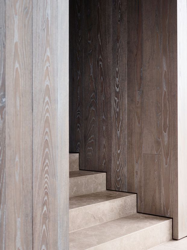 Close-up interior view of a staircase with broad beige stone or porcelain-clad treads and risers, framed by dark textured vertical timber wall panelling. A lighter timber-toned panel forms the foreground edge, creating a layered, refined stairwell composition.