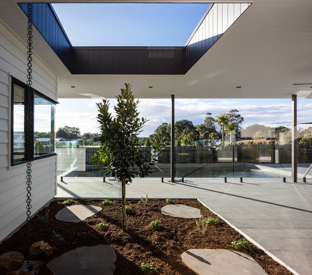 A contemporary covered outdoor courtyard or terrace beside a white weatherboard house, with a central planted garden bed, stepping stones, expansive grey tiled paving, slim dark posts, and a glazed pool area beyond. The roof forms a large open rectangular skylight that brings direct sunlight into the space. Suburban trees and neighbouring roofs are visible through the glass and open sides.