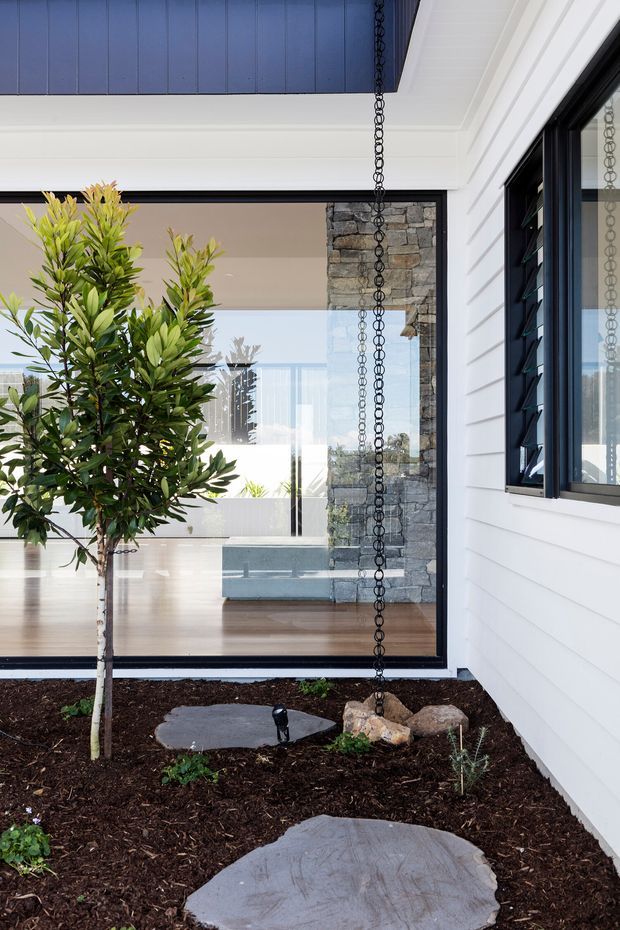 A contemporary house courtyard viewed from outdoors, with white horizontal weatherboard cladding, dark-framed glazing, a blue-painted soffit or ceiling, and a black rain chain descending into a landscaped garden bed. Large stepping stones lead toward full-height glass doors, through which a bright interior with timber flooring, a stone-clad structural column or fireplace, and reflective surfaces is visible. The composition highlights the connection between the landscaped courtyard and the interior.