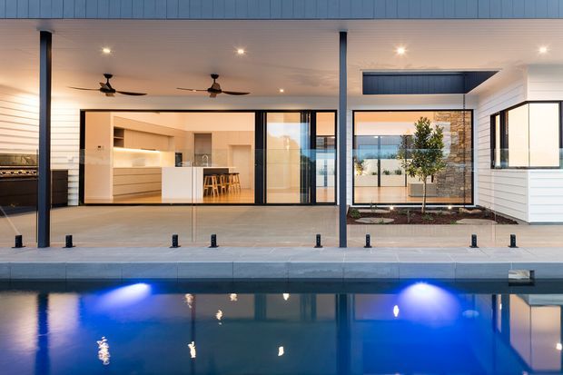 A contemporary single-storey residence viewed from the swimming pool, with a covered outdoor entertaining area, slim dark structural posts, ceiling fans, and expansive black-framed glazing opening onto a light-toned kitchen and living zone. White horizontal cladding, warm interior lighting, landscaped courtyard planting, and blue pool lighting create a refined indoor-outdoor setting.