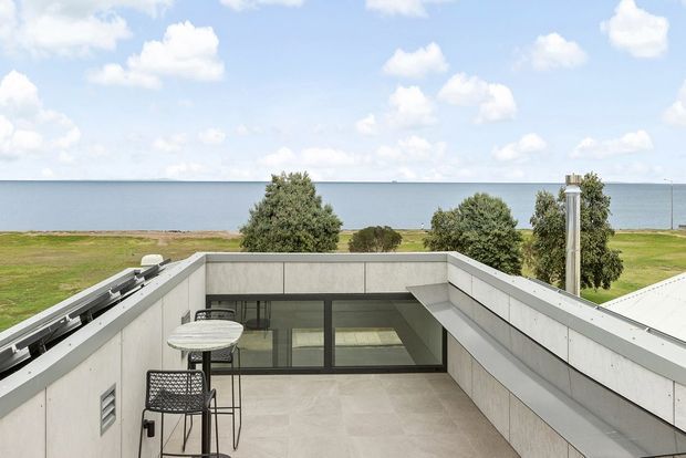 A rooftop terrace viewed from above, enclosed by low pale stone or concrete parapet walls. The terrace includes a small round table and bar stools, with large glazed doors opening into the building. Beyond the terrace is a flat coastal landscape and expansive body of water under a bright, partly cloudy sky. The image has a clean, modern architectural character.